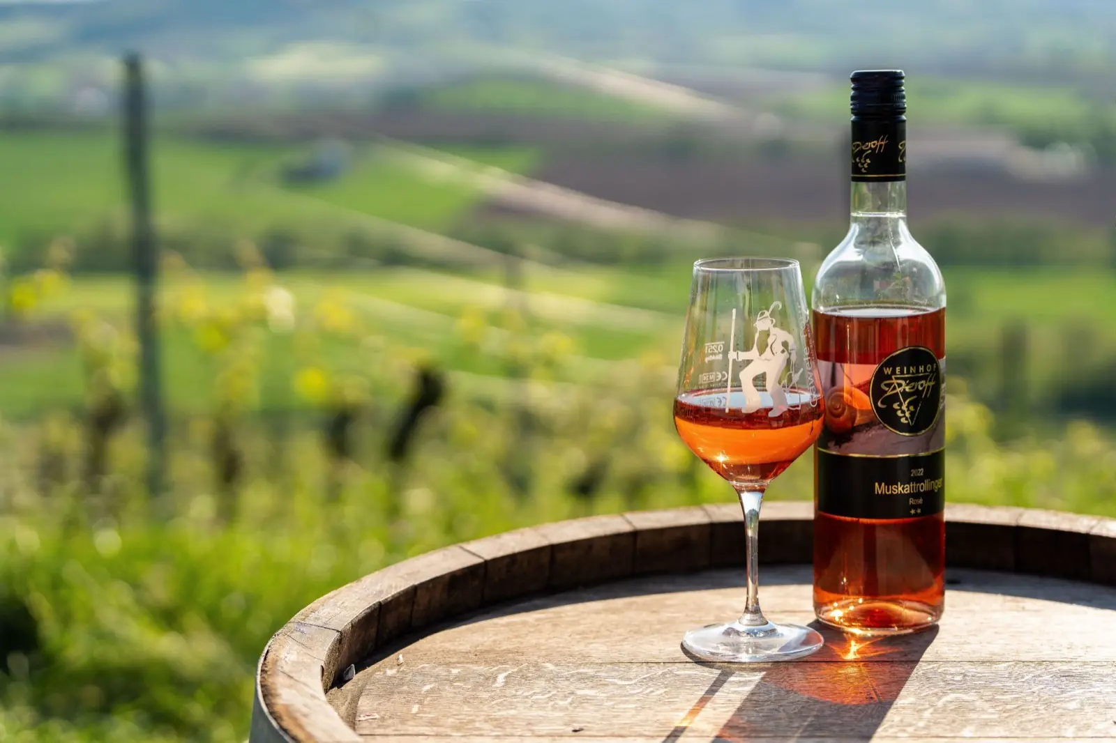 Eine Flasche Muskattrollinger Rosé und ein gefülltes Weinglas stehen auf einem Holzfass, dahinter die Weinberge von Michelbach am Wald.