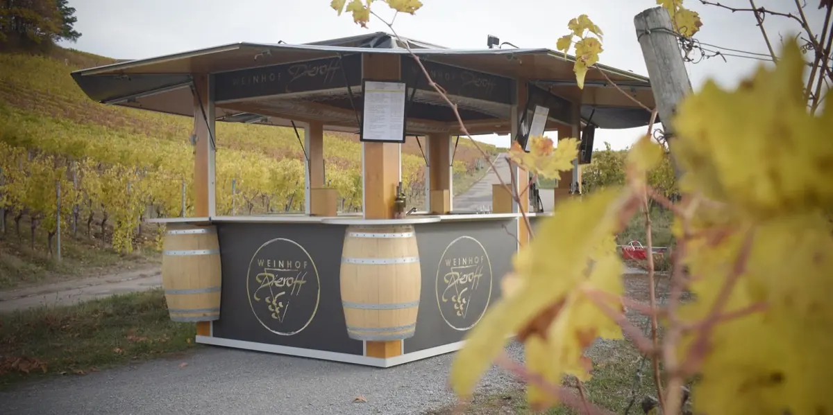 Der mobile Weinstand des Weinhofs Dieroff mit Holzdach und zwei Fässern, dahinter herbstlich gelb gefärbte Weinberge.